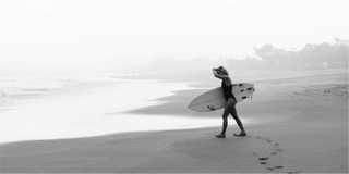 Eine Person, die mit einem Surfbrett am Strand spaziert, umgeben von natürlichen Elementen.